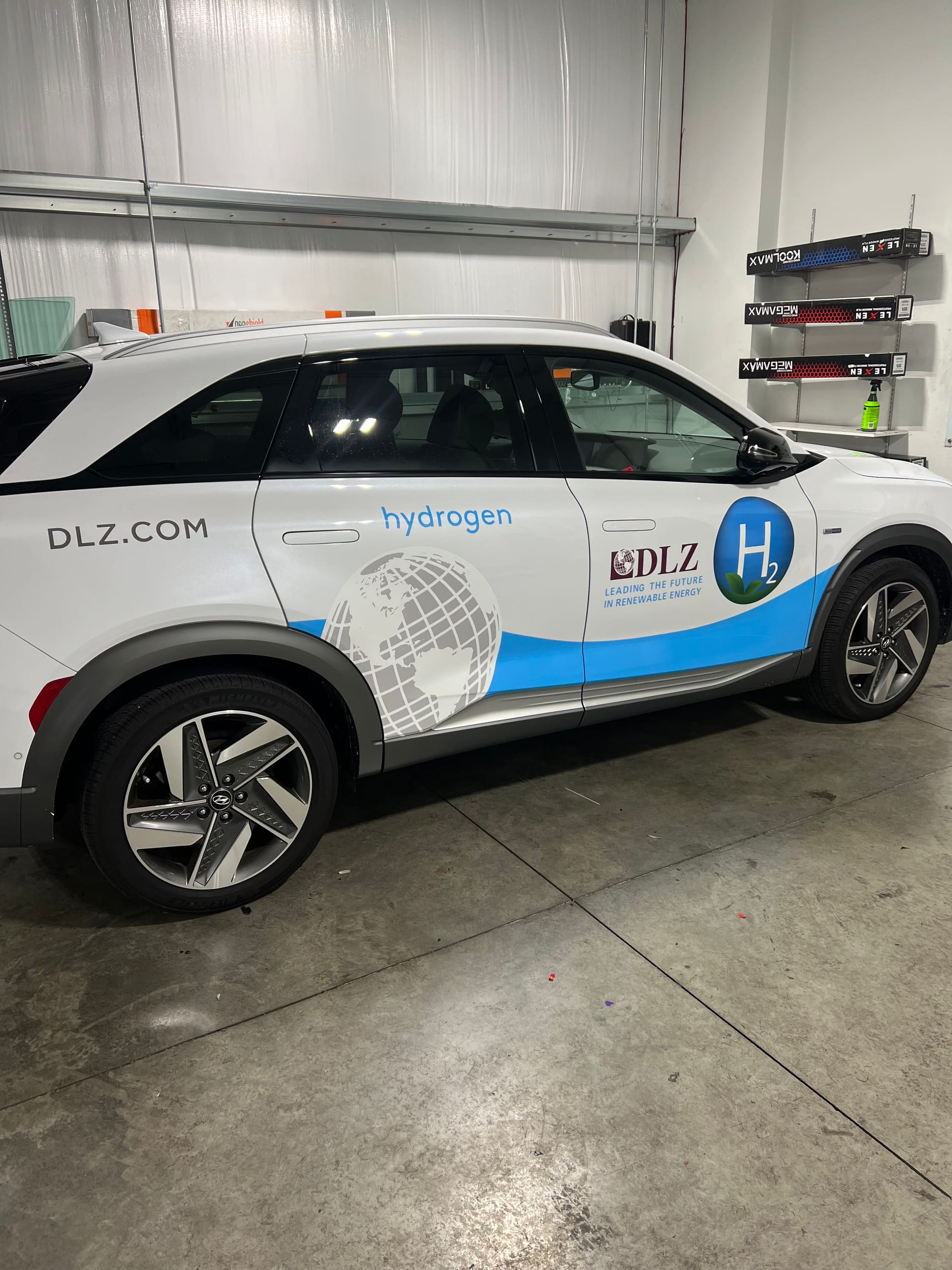 Hyundai Nexo hydrogen SUV in a white and blue DLZ partial wrap, photographed inside the shop bay with sealed concrete floor and boxed film rolls racked on the wall behind