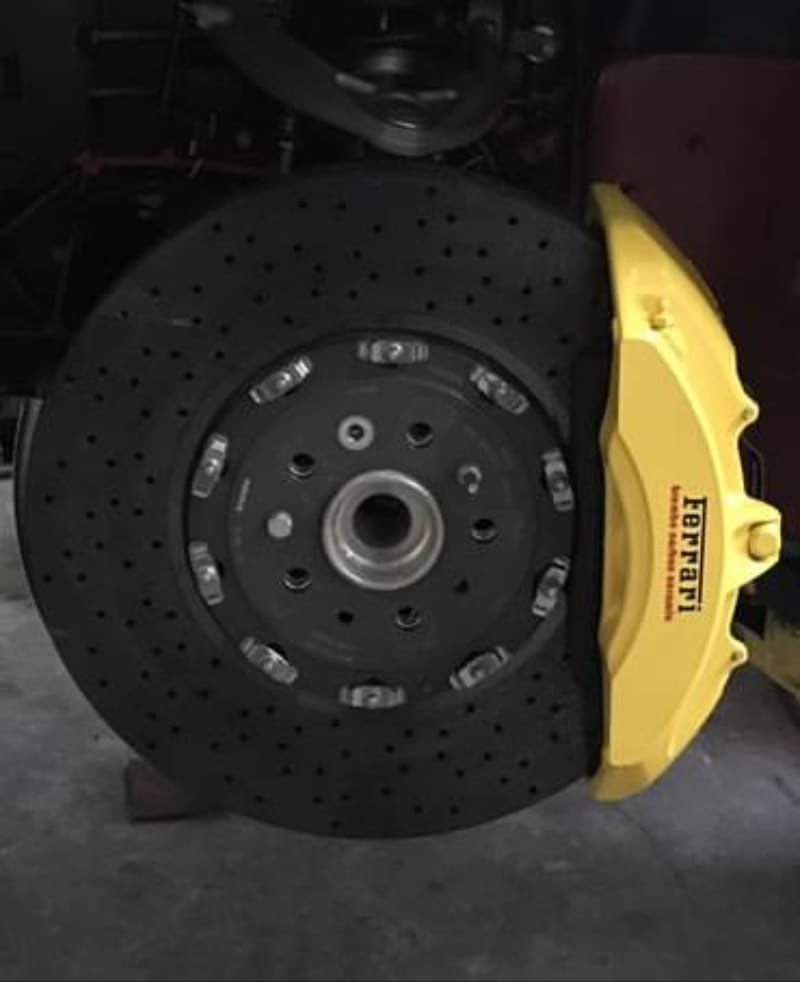 A Ferrari with the wheel removed, showing the yellow brake caliper against a drilled carbon-ceramic rotor, car raised in the workshop