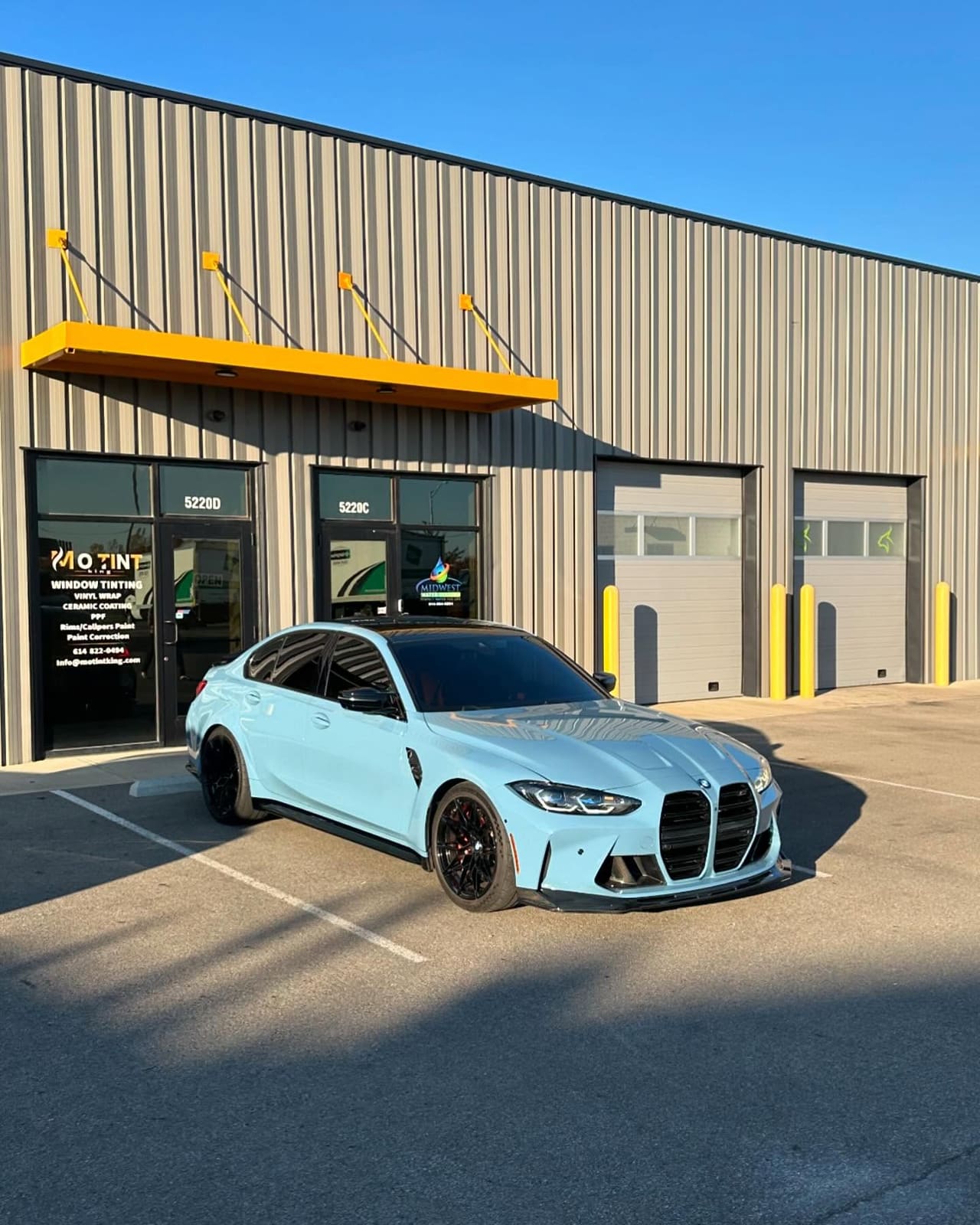 Light blue BMW M3 with black wheels, black roof and a black boot spoiler parked outside Mo Tint, the yellow awning above and units 5220D and 5220C legible on the glass beside the MO TINT decal listing window tinting, vinyl wrap, ceramic coating, PPF, rims and calipers paint and paint correction
