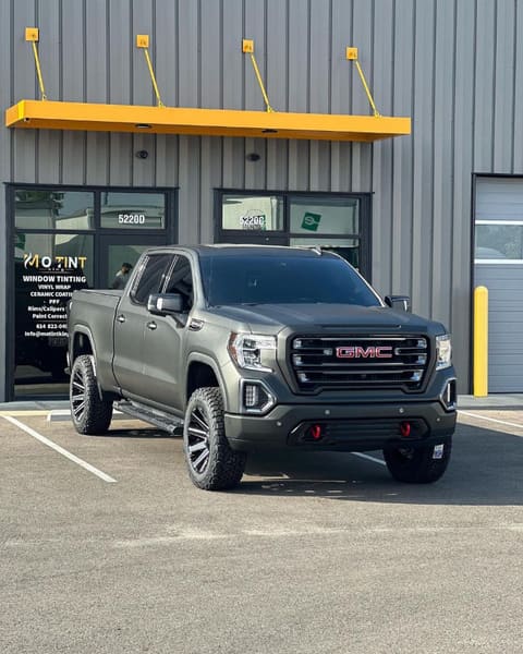 Grey GMC Sierra AT4 with tinted windows and red-painted calipers parked outside Mo Tint's unit 5220D, the MO TINT decal and service list visible on the glass behind it