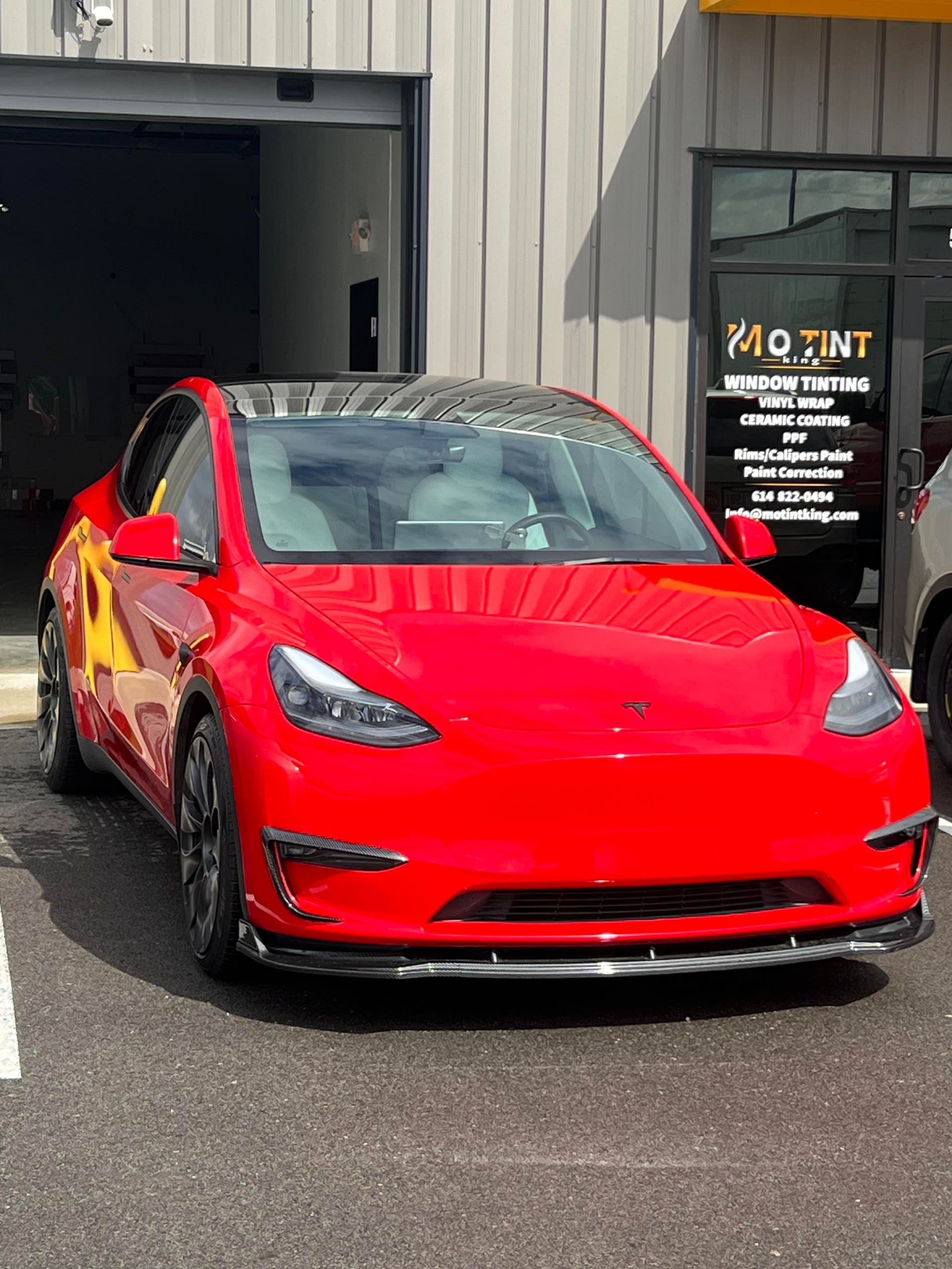 Red Tesla Model Y parked beside Mo Tint's open roller door, the MO TINT sign with service list and phone number on the wall behind