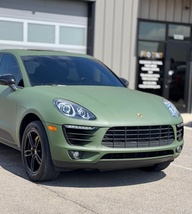Front end of a matte army green Porsche Macan beside the shop's roller door, with the wall-mounted MO TINT signboard at right