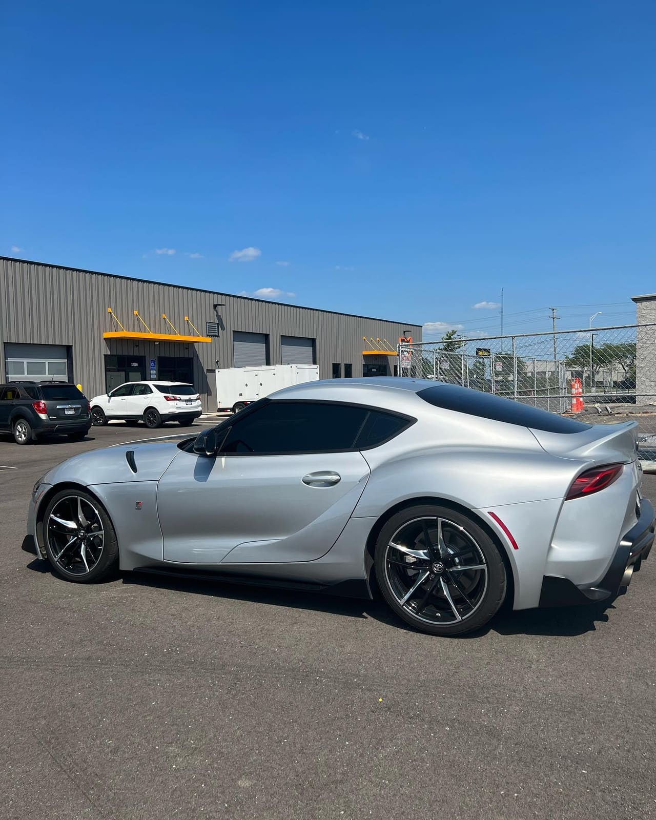 Rear three-quarter of a silver Toyota GR Supra with tinted windows, the shop's bay doors and yellow awning in the background