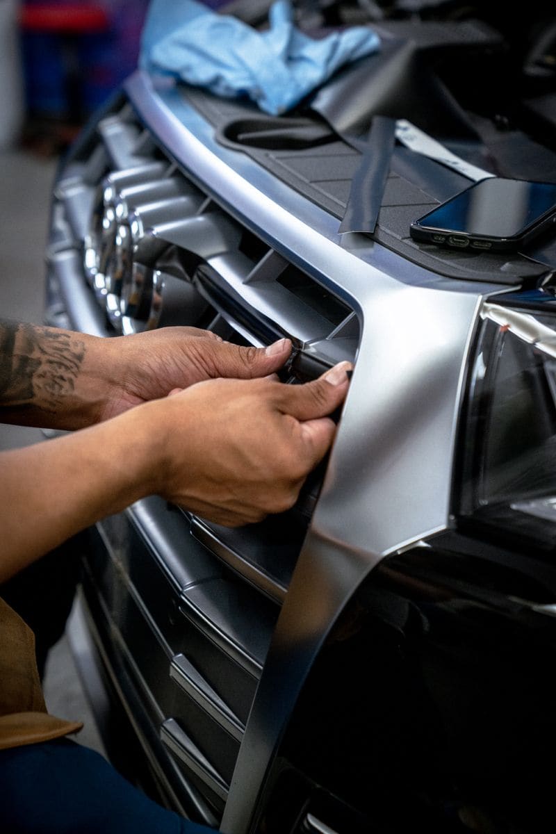 Two hands working a satin silver vinyl panel around the front bumper corner of a vehicle with a squeegee